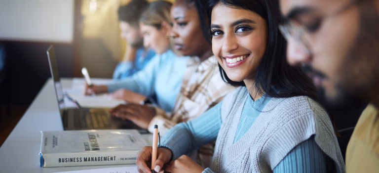 Business Mangement students of various ethnicities in a classroom with a book on the topic and taking notes, some on paper and on on a laptop.