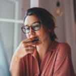 Image of a woman wearing glasses holding a pen staring into a computer screen.